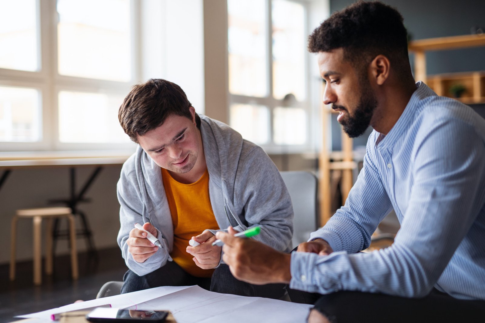 Young happy man with Down syndrome and his tutor studying indoors at school.
